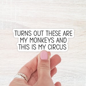 May include: A white sticker with black text that reads "TURNS OUT THESE ARE MY MONKEYS AND THIS IS MY CIRCUS." The sticker is rectangular with rounded corners. A hand with a pink painted nail is holding the sticker.