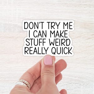 May include: A white sticker with black text that reads "DON'T TRY ME I CAN MAKE STUFF WEIRD REALLY QUICK." The sticker is held by a hand with pink nail polish and rings.