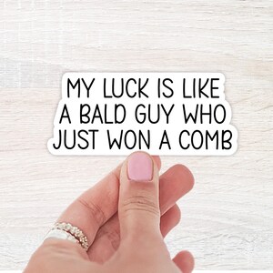 May include: A white sticker with black text that reads, "MY LUCK IS LIKE A BALD GUY WHO JUST WON A COMB." The sticker is held by a hand with a pink painted nail and a silver ring.