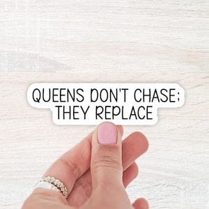 May include: A white sticker with the text "QUEENS DON'T CHASE; THEY REPLACE" in black, held by a hand with pink nail polish and rings. The sticker is on a light wood grain surface.
