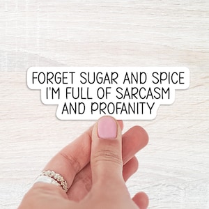 May include: A white sticker with black text that reads "FORGET SUGAR AND SPICE I'M FULL OF SARCASM AND PROFANITY." The sticker is held by a hand with a pink painted fingernail and a silver ring.
