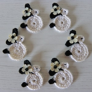 Può includere: Sei pecorelle all'uncinetto bianche con dettagli neri e un fiore giallo su ciascuna. Le pecorelle sono disposte in cerchio su una superficie bianca.