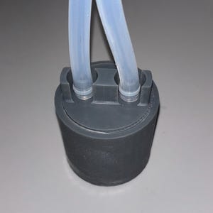 May include: A dark gray cylindrical component with two clear, flexible tubes extending upwards. The tubes are connected to the top of the cylinder. The component appears to be made of plastic or a similar material.