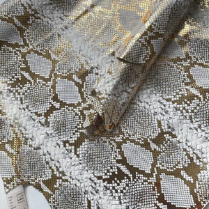 May include: Close-up of a white and gold snakeskin fabric with a textured pattern. The fabric is folded in half, showing the inside and outside of the material.