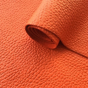 May include: Close-up of a bright orange leather material with a textured surface. The leather is rolled up, showing the thickness and grain.