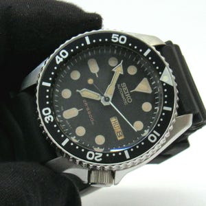 Könnte beinhalten: Eine Seiko Automatikuhr mit schwarzem Zifferblatt, leuchtenden runden Stundenmarkierungen und einer schwarzen drehbaren Lünette mit weißen Ziffern. Die Uhr verfügt über eine Tages-Datums-Anzeige auf der Drei-Uhr-Position und ein schwarzes Kautschukarmband.