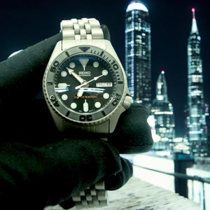 May include: A silver Seiko automatic dive watch with a black dial, luminous markers, and a date display. The watch has a stainless steel bracelet and a rotating bezel. The background features city skyscrapers at night.