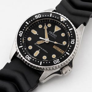 May include: A Seiko automatic diver's watch with a black dial, luminous hour markers, and hands. The watch features a black rotating bezel with white numerals, a day-date window at the three o'clock position, and a black rubber strap.