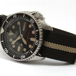 May include: A stainless steel Seiko automatic diver watch with a black dial, luminous circular hour markers, and a day-date window. The watch features a black rotating bezel with white numerals and is attached to a black and tan striped nylon strap.