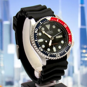 May include: A Seiko automatic wristwatch with a black dial, stainless steel case, and black rubber strap. The watch has a rotating bezel with red and blue detailing. The watch face shows the day and date.