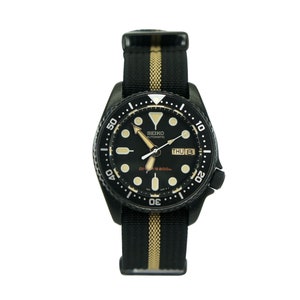 May include: Black Seiko automatic dive watch with a black and gold striped nylon strap. The watch face has a black dial with white markings and a date window.