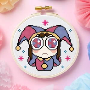 Può includere: Un telaio da ricamo a punto croce con un personaggio di giullare pixelato con una combinazione di colori rosa e blu. Il giullare ha grandi occhi rotondi e un'espressione triste. Lo sfondo è rosa con decorazioni di carta colorate.