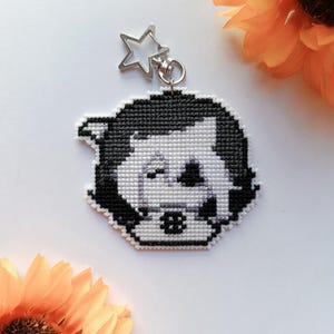 May include: A black and white cross-stitch keychain featuring a stylized character face with a winking eye and a small square detail on the chin. The embroidered piece is attached to a silver-colored metal star-shaped clasp and ring.
