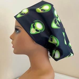 May include: Black surgical scrub cap featuring a repeating pattern of green alien faces. The alien faces have large black eyes and are outlined in white. The cap is made of a soft, flexible material and is designed to fit comfortably.
