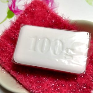 Pode incluir: Uma barra de sabão branca com o texto "100g" impresso nela. O sabão está sobre uma superfície felpuda rosa e branca.