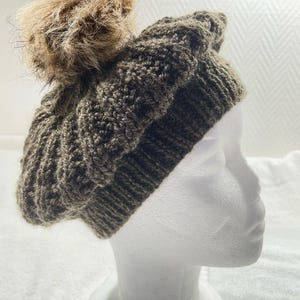 Può includere: Un basco lavorato a maglia verde oliva con un soffice pompon marrone chiaro. Il cappello è esposto su una testa di manichino bianca, mettendo in risalto la consistenza e la forma dell'accessorio invernale. Il basco è un copricapo elegante e caldo.
