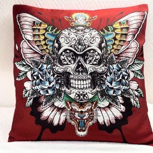 Può includere: Un cuscino quadrato decorativo con sfondo rosso. Il cuscino presenta un design dettagliato di un teschio con ali di farfalla, rose blu e una faccia di tigre. Il teschio è bianco con dettagli neri.
