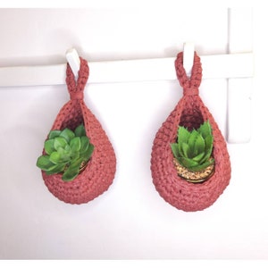 May include: Two terracotta-colored crocheted hanging planters with green succulent plants inside. The planters are hanging from a white shelf.