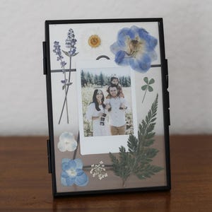 May include: A rectangular black metal floating frame containing a central instant photograph of a family of three. The glass is decorated with pressed blue and white flowers, a green fern leaf, and a small four-leaf clover arrangement.