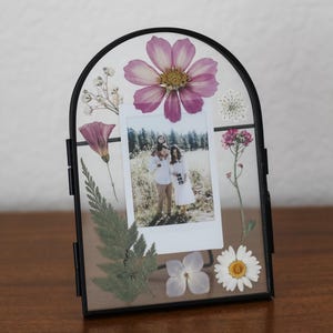 May include: An arched black metal floating frame containing a vertical rectangular photograph of a family in a field. The glass is decorated with pressed dried flowers including a large pink cosmos, a white daisy, ferns, and small white blossoms.