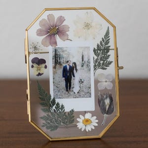 May include: A gold-framed photo holder with an octagonal shape. The frame contains a photo of a couple and is decorated with pressed flowers and greenery. The flowers include a purple cosmos, white daisy, and a black tulip.