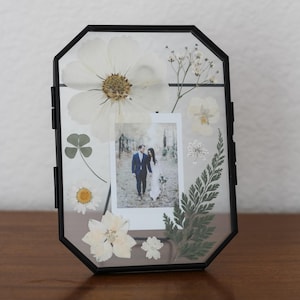 May include: An octagonal black metal floating frame containing a small rectangular photograph of a couple walking in a forest, surrounded by pressed white flowers, green fern leaves, and a four-leaf clover arranged between two clear glass panes.