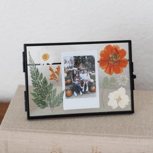 May include: A rectangular black metal floating frame containing a central instant photograph of a family sitting among pumpkins, surrounded by pressed botanical elements including a green fern, a white daisy, and an orange flower.