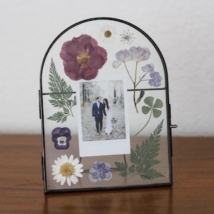 May include: An arched glass picture frame with a thin black metal border containing pressed dried flowers in shades of purple, white, and green. A small rectangular photograph of a couple is centered between the botanical elements.