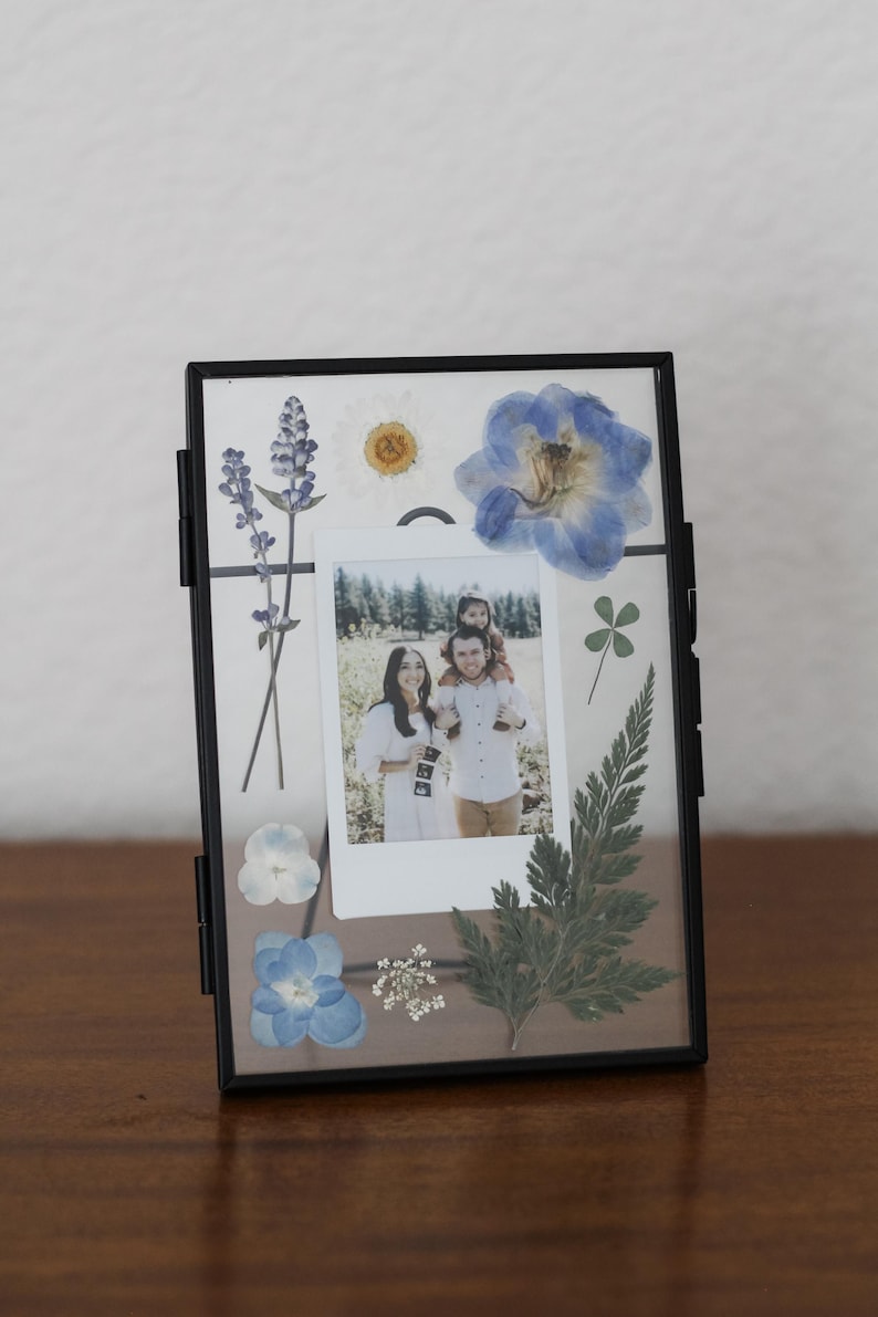 May include: A rectangular black metal floating frame containing a central instant photograph of a family of three. The glass is decorated with pressed blue and white flowers, a green fern leaf, and a small four-leaf clover arrangement.