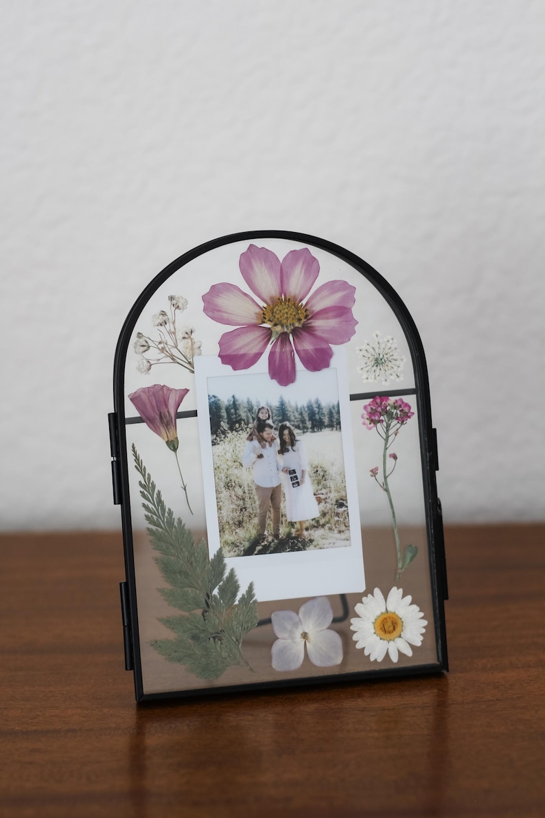 May include: An arched black metal floating frame containing a vertical rectangular photograph of a family in a field. The glass is decorated with pressed dried flowers including a large pink cosmos, a white daisy, ferns, and small white blossoms.