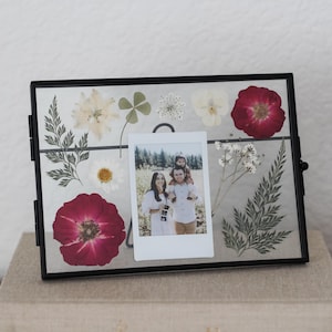 May include: A rectangular black metal floating frame containing pressed dried flowers and greenery, including red roses, white daisies, and ferns. A small vertical instant film photograph of two people is centered within the glass display.