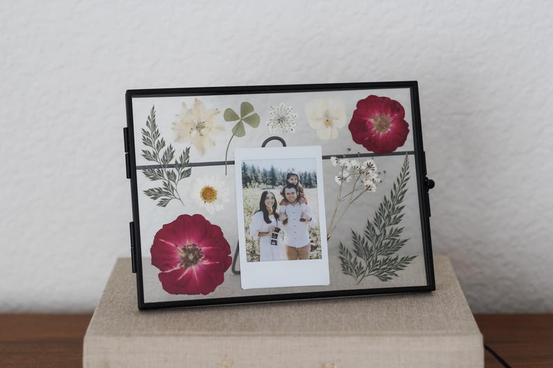 May include: A rectangular black metal floating frame containing pressed dried flowers and greenery, including red roses, white daisies, and ferns. A small vertical instant film photograph of two people is centered within the glass display.