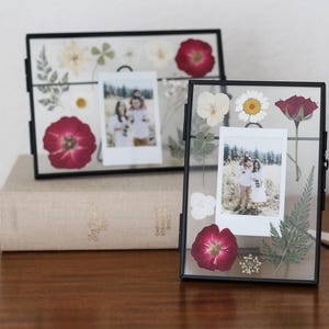 May include: Two rectangular floating picture frames with thin black metal borders. Each frame contains pressed dried flowers in shades of red, white, and green, surrounding a central vertical instant film photograph of two people in a field.