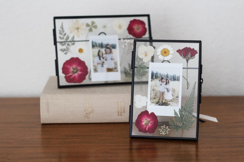 May include: Two rectangular floating picture frames with thin black metal borders. Each frame contains pressed dried flowers in shades of red, white, and green, surrounding a central vertical instant film photograph of two people in a field.