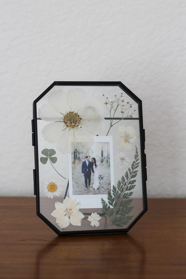 May include: An octagonal black metal floating frame containing a small rectangular photograph of a couple walking in a forest, surrounded by pressed white flowers, green fern leaves, and a four-leaf clover arranged between two clear glass panes.