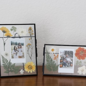 May include: Two rectangular black metal floating frames containing pressed dried flowers and foliage in shades of yellow, orange, white, and green. Each frame holds a central vertical or horizontal instant film photograph of people.