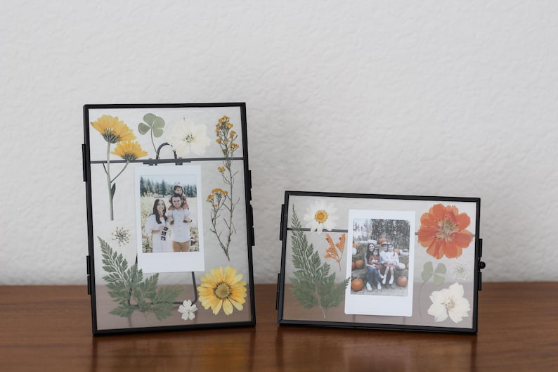 May include: Two rectangular black metal floating frames containing pressed dried flowers and foliage in shades of yellow, orange, white, and green. Each frame holds a central vertical or horizontal instant film photograph of people.