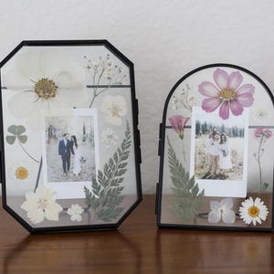 May include: Two black metal floating frames containing pressed flowers and photographs. The left frame is octagonal with white flowers and ferns. The right frame is arched with pink and white flowers and ferns. Both hold small square photos.
