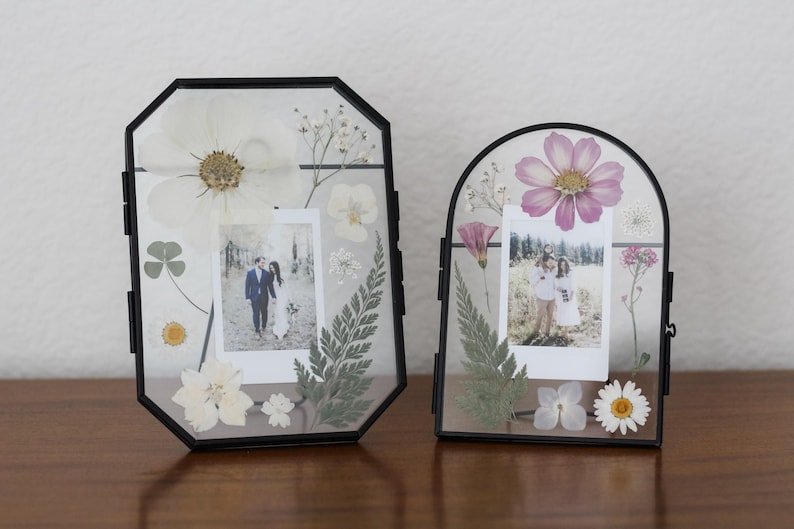 May include: Two black metal floating frames containing pressed flowers and photographs. The left frame is octagonal with white flowers and ferns. The right frame is arched with pink and white flowers and ferns. Both hold small square photos.