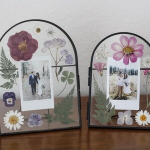 May include: Two arched glass frames with black metal borders containing pressed flowers and foliage surrounding a central photograph. The left frame features a dark purple flower and ferns, while the right frame displays a pink flower and greenery.