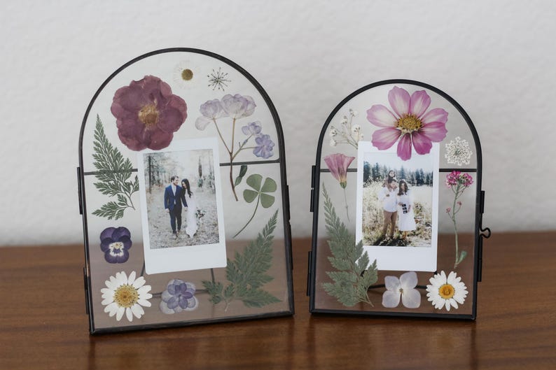 May include: Two arched glass frames with black metal borders containing pressed flowers and foliage surrounding a central photograph. The left frame features a dark purple flower and ferns, while the right frame displays a pink flower and greenery.