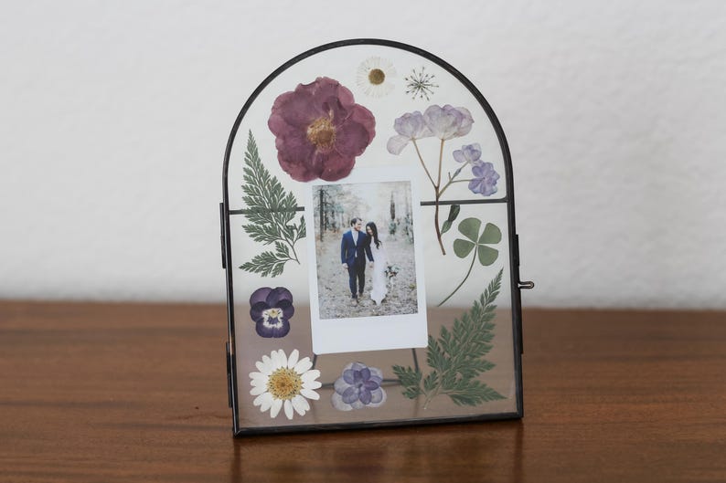 May include: An arched glass picture frame with a thin black metal border containing pressed dried flowers in shades of purple, white, and green. A small rectangular photograph of a couple is centered between the botanical elements.