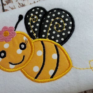 May include: A yellow and black embroidered bee with white polka dots. The bee has a pink flower on its head and a yellow tail.