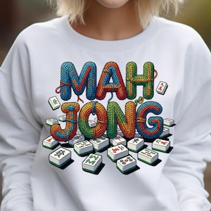 May include: White sweatshirt featuring the word "MAH JONG" in colorful, knitted-style letters. The design includes scattered Mahjong tiles with Chinese characters. The sweatshirt is a casual garment.