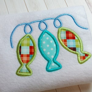 May include: Embroidered design featuring three fish in shades of green, blue, and red on a white background. The fish are connected by a blue, swirled line. The fish have different patterns, including plaid and polka dots.