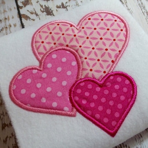Puede incluir: Un cuadrado blanco con tres corazones de tela rosa. Los corazones tienen diferentes patrones: lunares y un diseño floral. Cada corazón está delineado con un hilo rosa más oscuro.