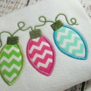 May include: Embroidered Christmas light design featuring three colorful bulbs: green, pink, and blue. Each bulb has a white zigzag pattern and a green stem. The design is on a white fabric.