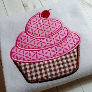 Puede incluir: Una aplicación de cupcake de tela rosa y marrón con una cereza roja en la parte superior. El cupcake está decorado con un patrón floral rosa y blanco y un patrón a cuadros marrón y blanco.