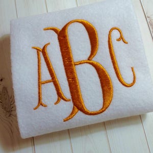 Peut inclure: Un tissu blanc avec un monogramme brodé en or des lettres "ABC" dans une police d'écriture cursive.