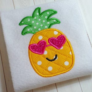 May include: Embroidered pineapple with a green leafy top, yellow body with white polka dots, and pink heart-shaped sunglasses. The pineapple has a smiling face.